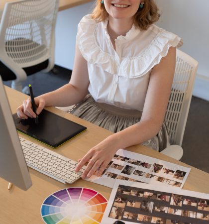 Portrait of young Caucasian female graphic designer working on graphics tablet and computer at desk in the office. She is smiling and looking at camera